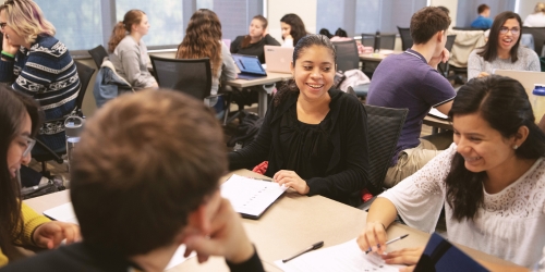 Students collaborate in small groups around tables in a classroom, talking and taking notes during a group activity.