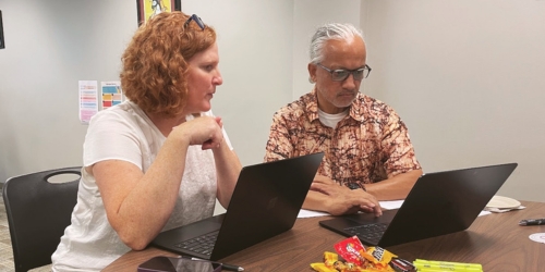 Two coworkers sit at a table with laptops, collaborating on a task in an office setting.