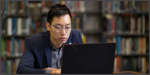 Young man wearing glasses works on a laptop at a table in a library, with bookshelves blurred in the background.