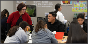 Instructor in a red cardigan interacts with smiling students in a classroom.