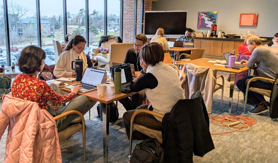 People working on laptops around tables in a lounge-like workspace with large windows and a coffee counter in the background.