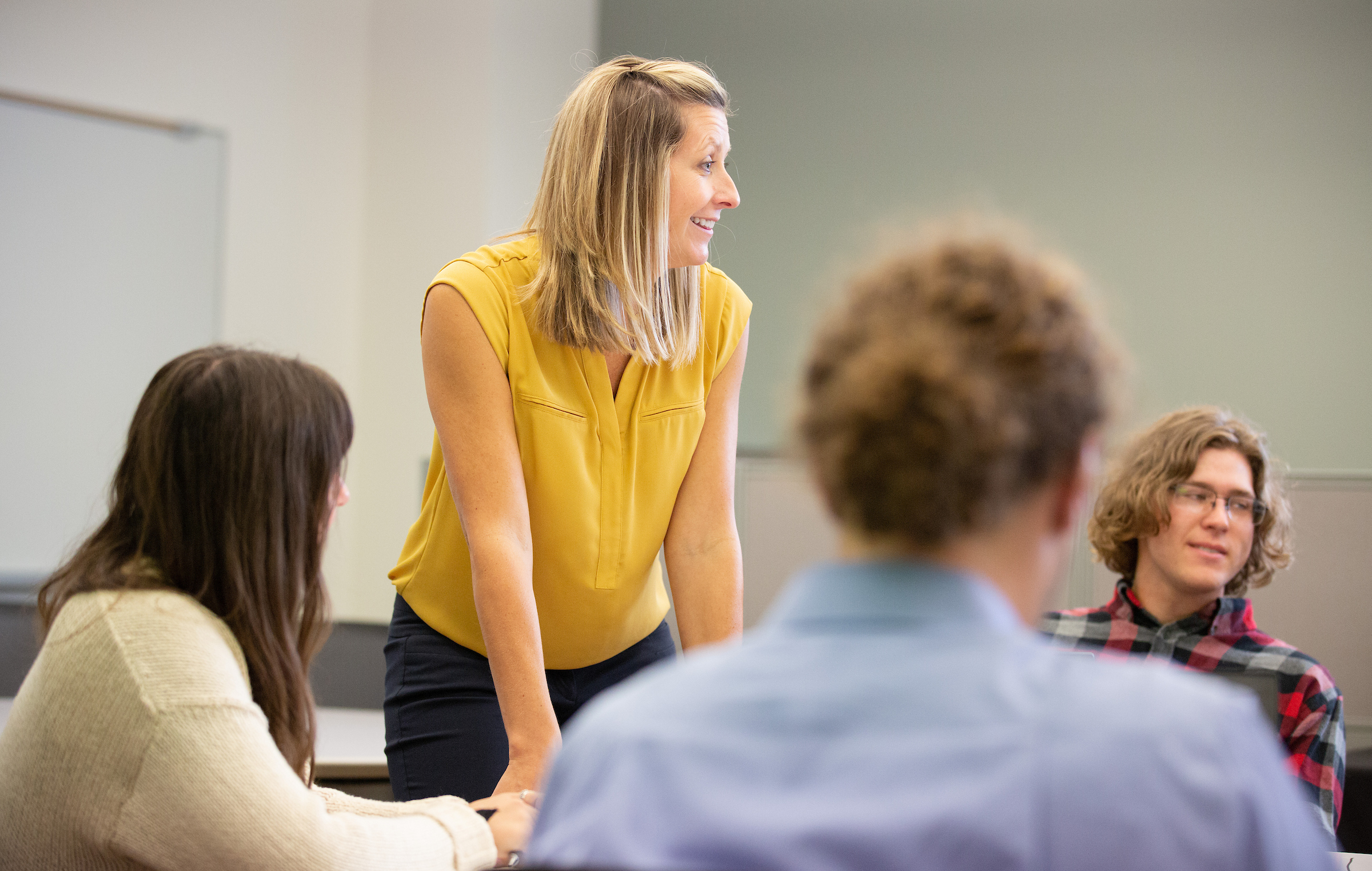 a female teacher in a yellow blouse leans on a desk as she has a discussion with students