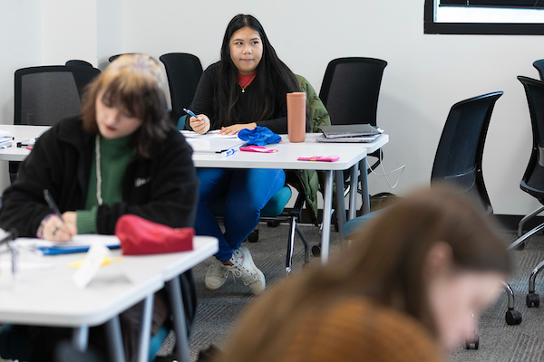 a female student of color sits at a desk in front of an open book while listening to the intructor