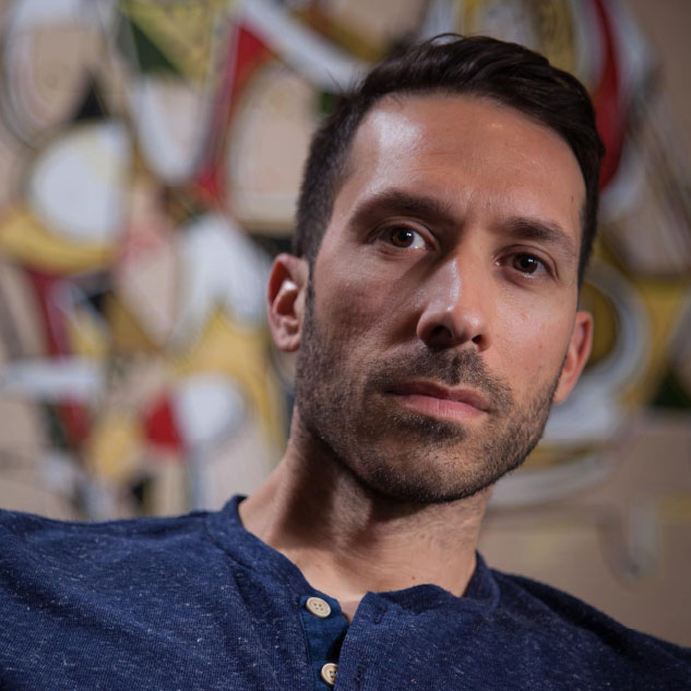 Man with short dark hair and trimmed beard wearing a navy blue henley shirt, gazing calmly at the camera with an abstract, colorful mural in the background.