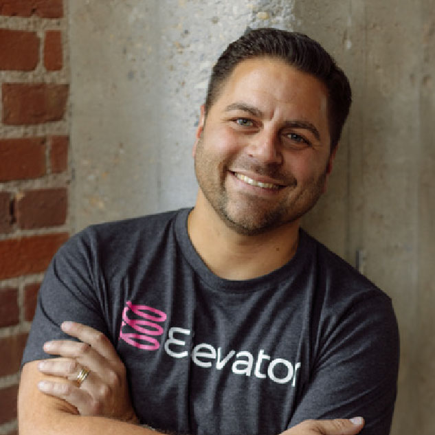 Smiling man with short dark hair and trimmed beard wearing a dark gray 'Elevator' T-shirt, standing with arms crossed in front of a concrete and brick wall.