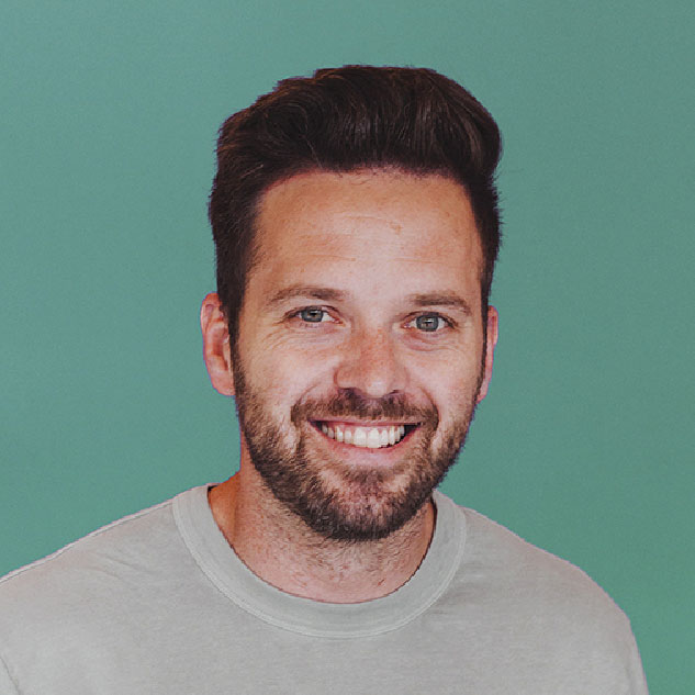 Smiling man with neatly styled dark hair and beard wearing a light gray T-shirt, standing in front of a solid teal background.