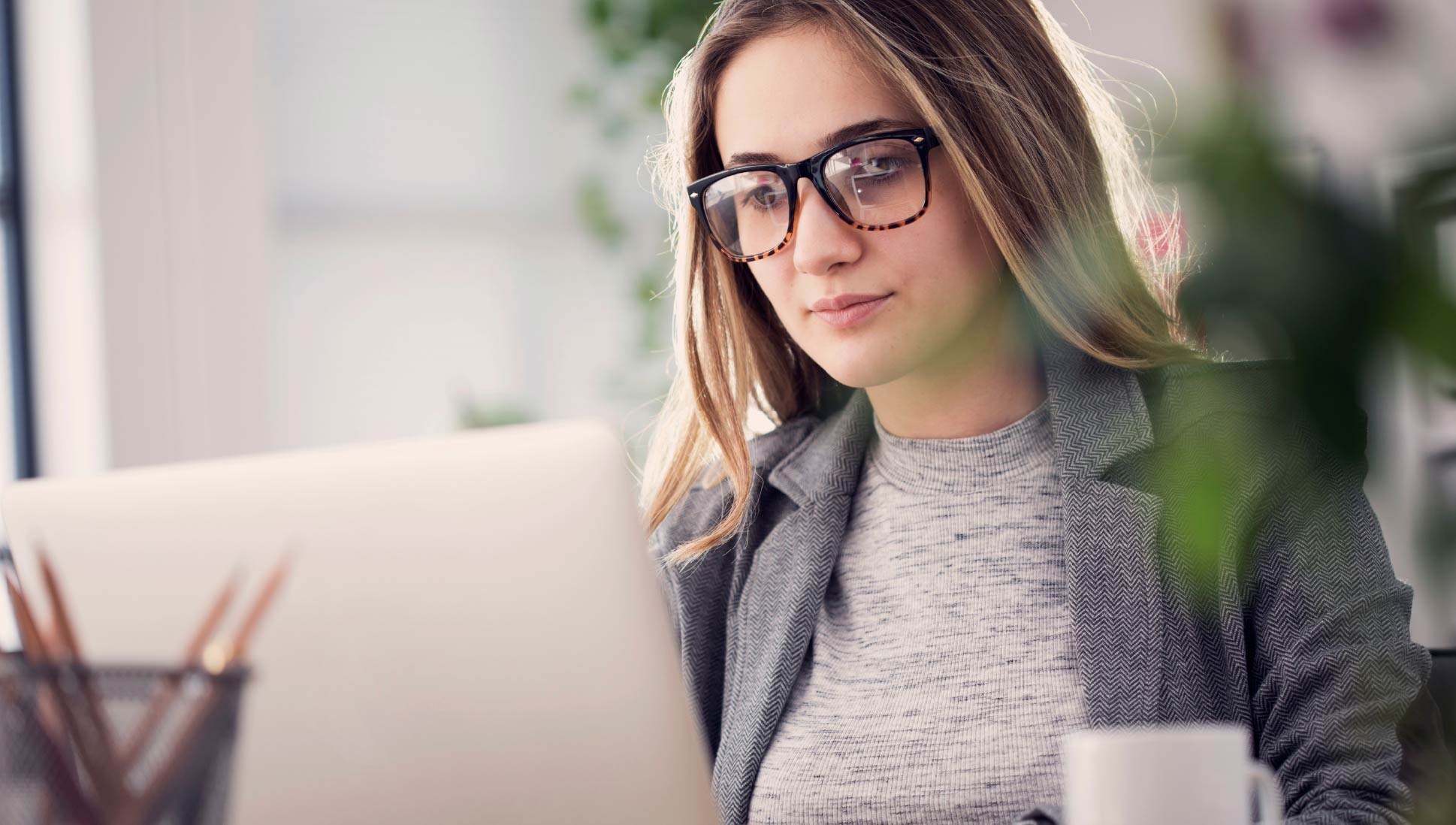 Woman wearing glasses works on a laptop at a desk in a bright indoor workspace.