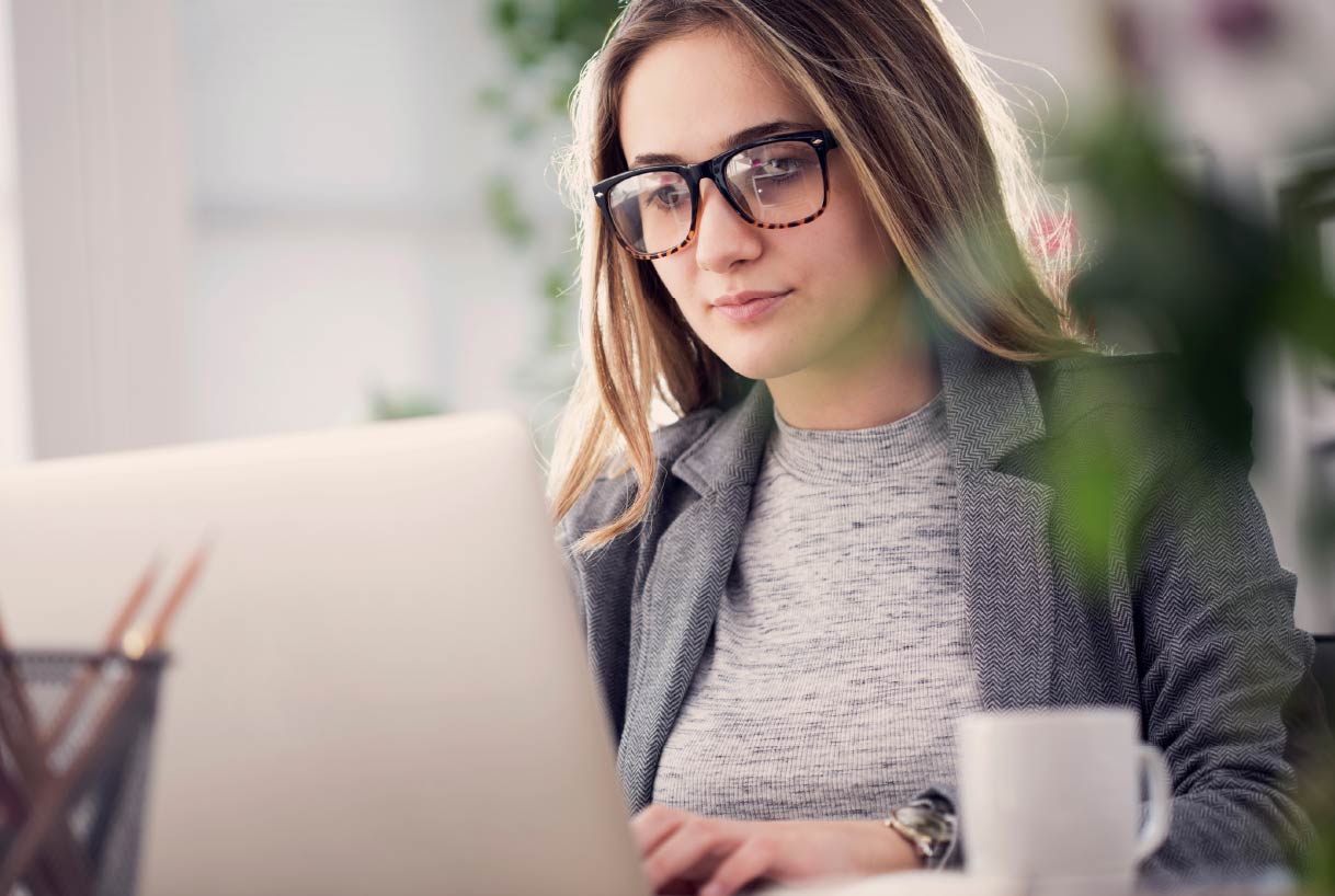 Woman wearing glasses works on a laptop at a desk in a bright indoor workspace.