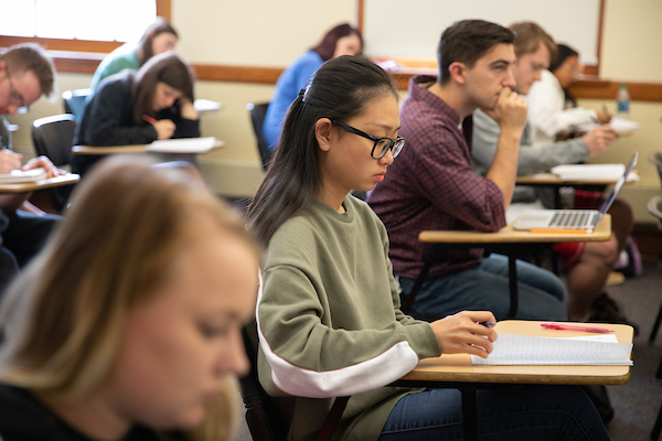 A college classroom with students seated at desks, listening and taking notes. In the center, a student wearing glasses and a green sweatshirt writes in a notebook, while other students around them focus on their notebooks or laptops.