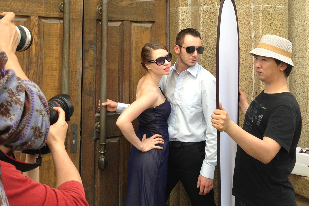 Michael Blakely and a woman, both in sunglasses, model for two photographers in front of a wooden door