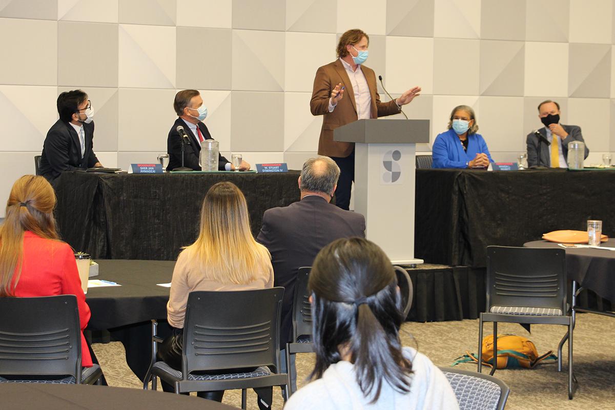 Michael Jung stands behind the podium with panelists seated to his left and right. Guests sit at tables in front of the stage listening.