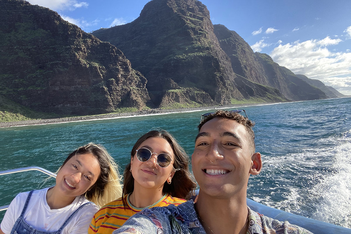 Three students smile in a boat over the water with a lush hillside in the background in an island setting.