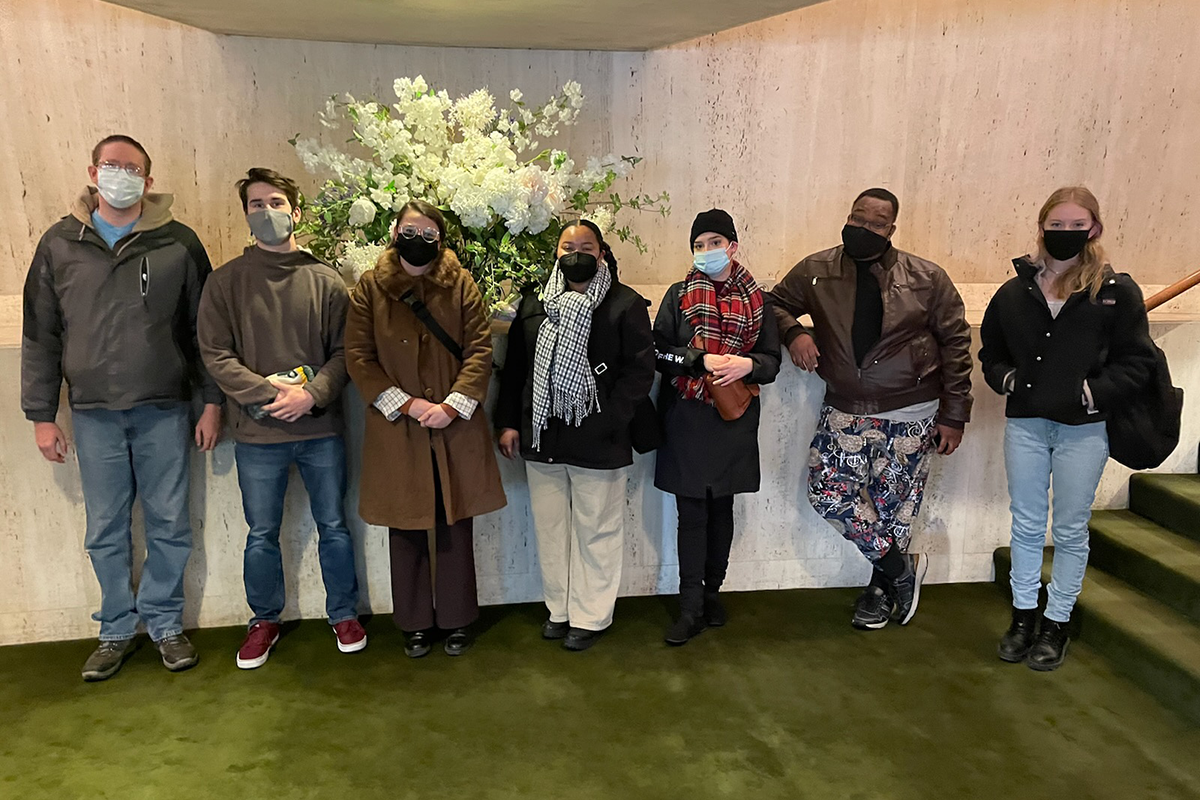 7 students stand in a lobby with a floral display behind them.