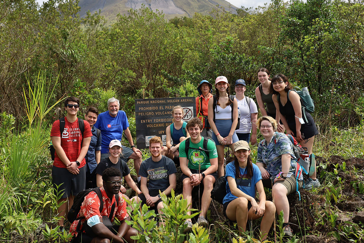 Group of students stand around the park sign for Arenal Volcano, with the volcano's base visible in the background.