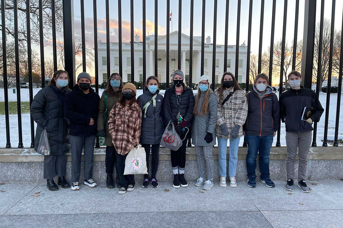 a group of masked students stand in front of a black metal fence, with The White House visible behind.