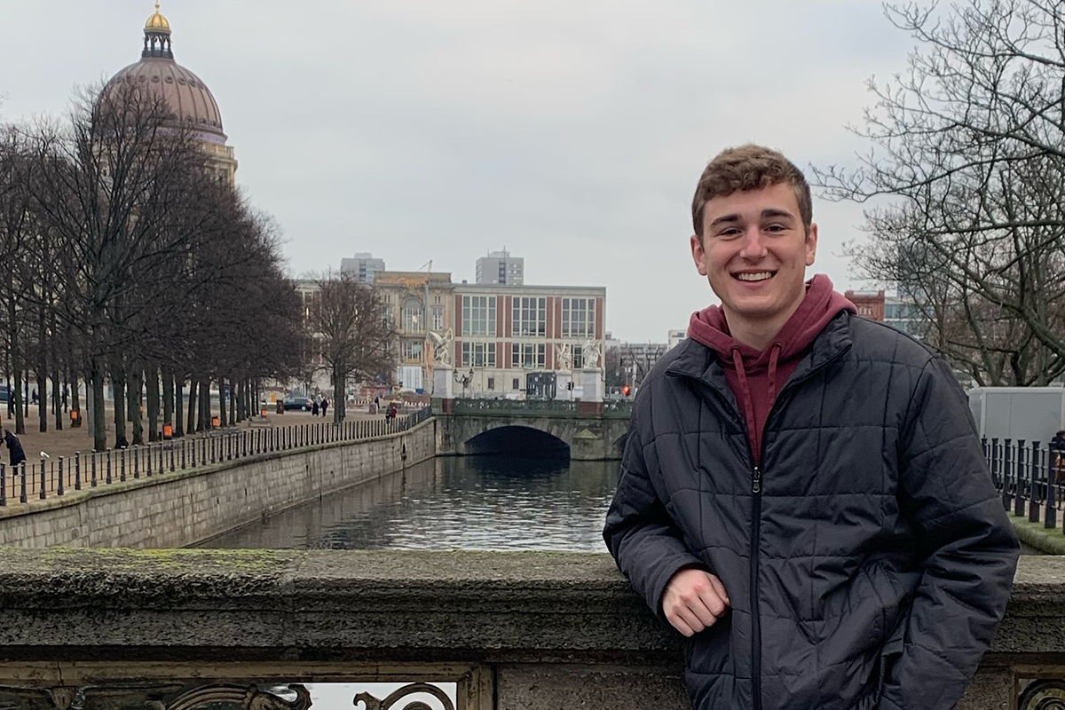 Student poses against a bridge railing in Berlin Germany, with a river and buildings in the background.