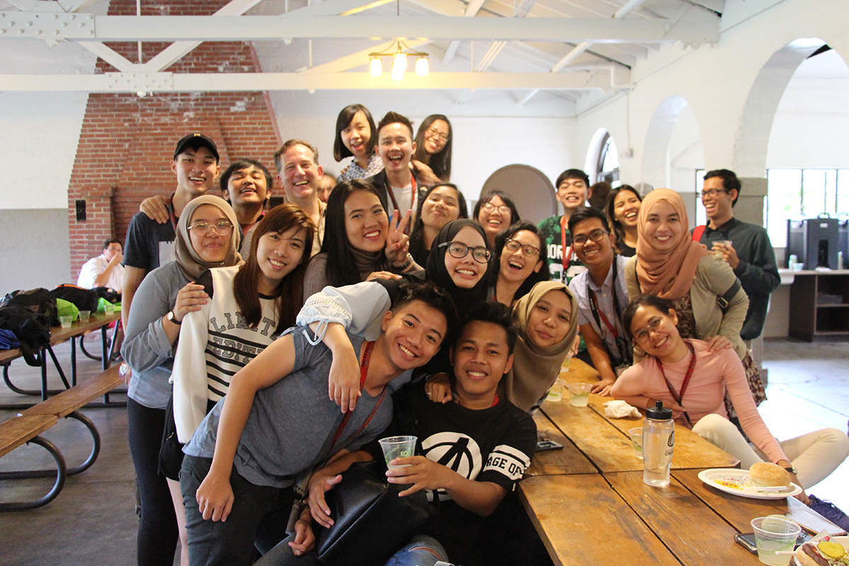 International students and visitors pose together in the Elmwood Park Pavilion around an indoor picnic table.