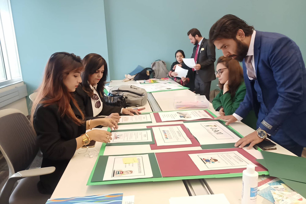 A group of teachers, some sitting some standing, work on presentation boards on the tables between them.