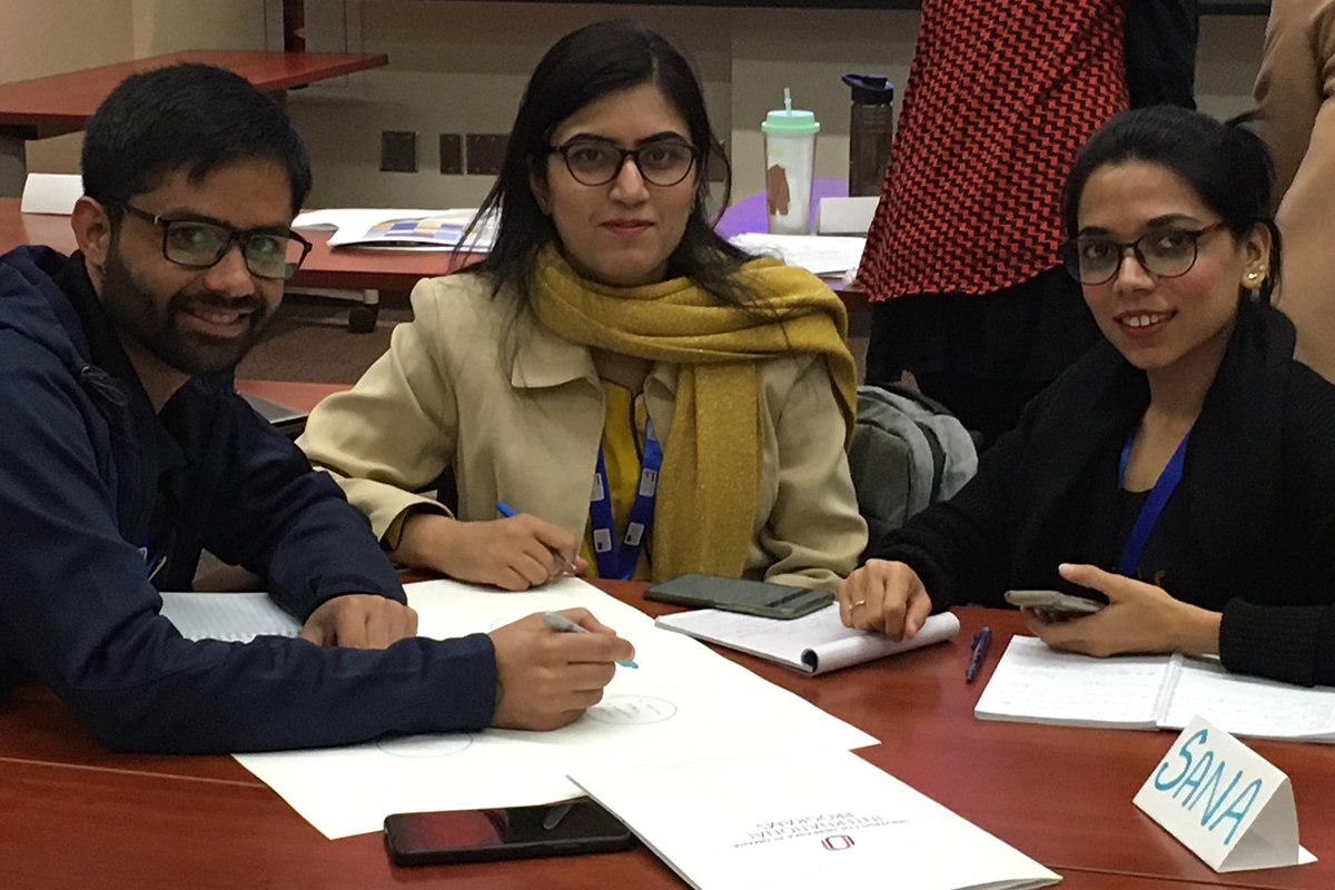 Three Pakistani English teachers sit at a desk together, with papers and folders in front of them.