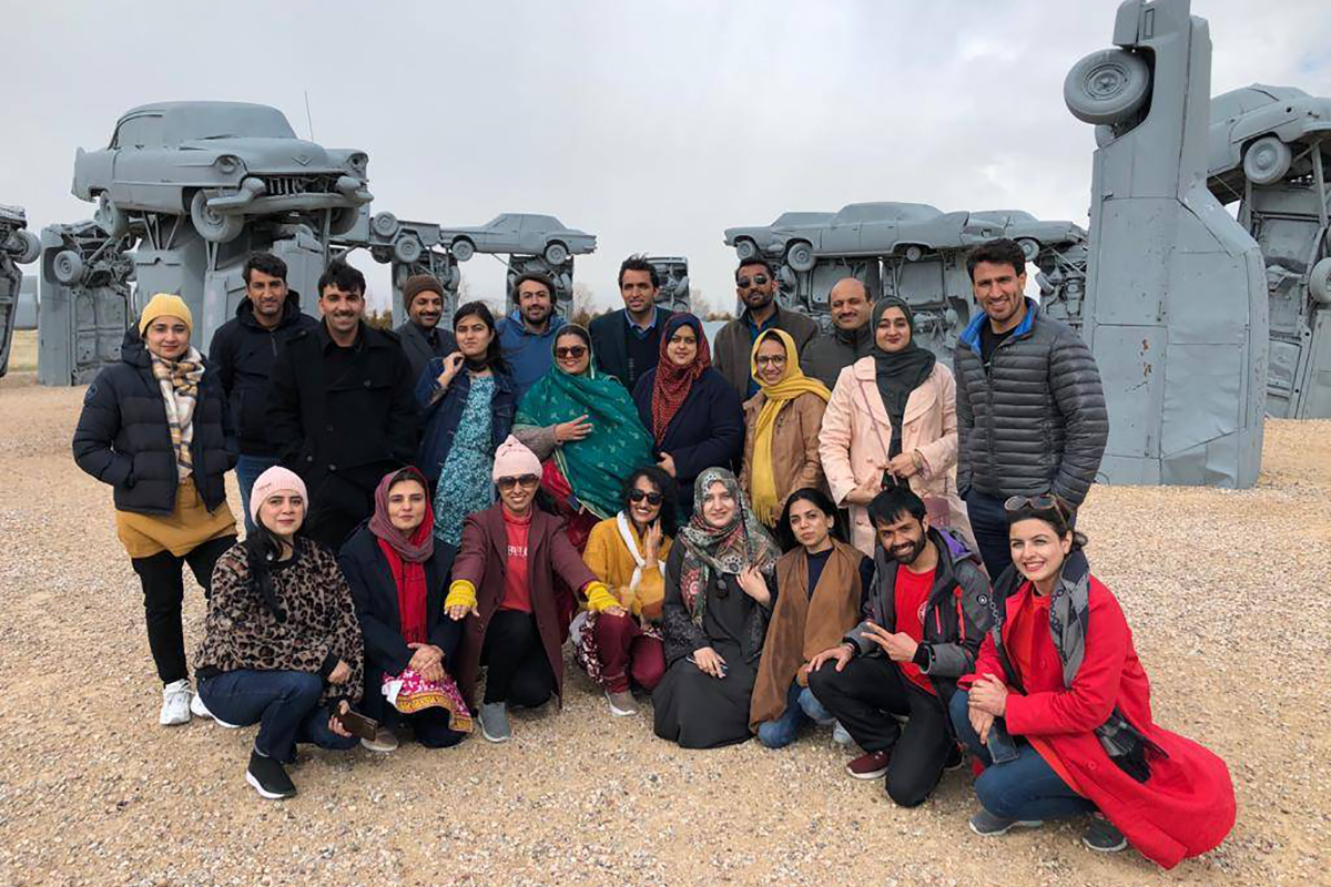 A group of teachers from Pakistan post in front of carhenge, a replica of stonehenge made out of cars painted grey.