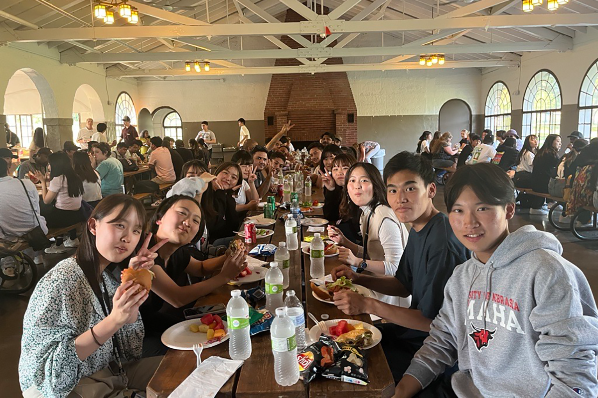 College students sit around a picnic table inside a park pavilion, eating hamburgers