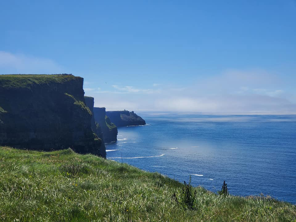 The sharp line of the rocky cliffs of Moher with green grassy tops appear on the left side of the image, while blue sky and ocean appear on the right.