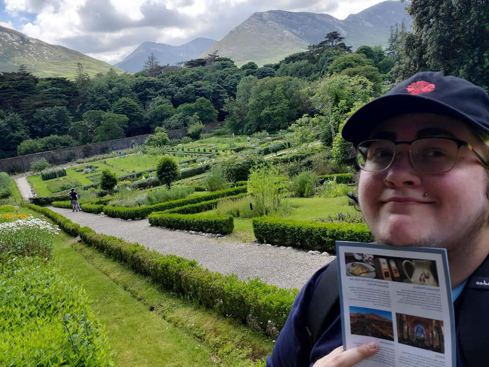 Young man with a baseball cap on snaps a selfie in front of a bright green garden with hedges. Trees and mountains appear in the distance.