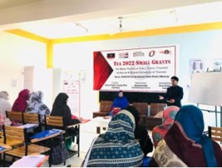 Teacher trainer stands at the front of a classroom with a presentation to his right. Pakistani female school teachers sit facing him.