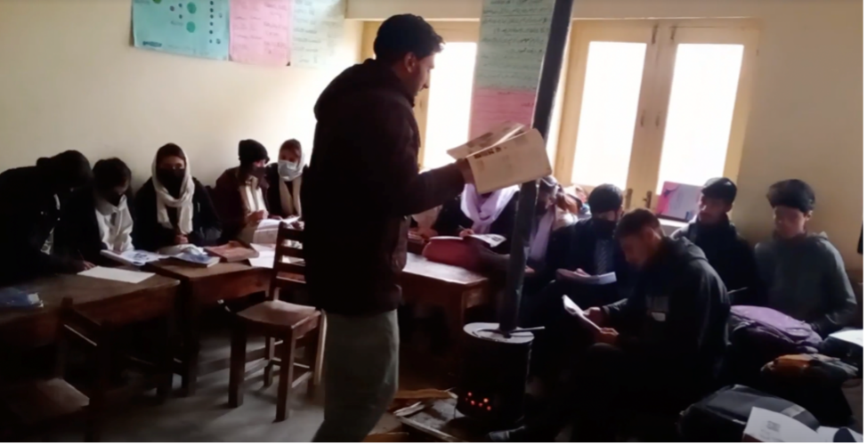 A male teacher stands in a classroom surrounded by teenagers at their desks.
