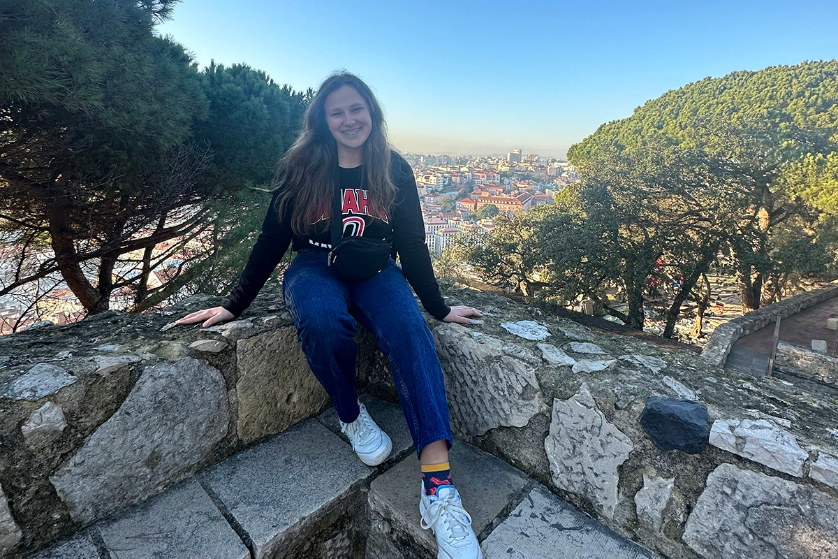 UNO student Ava Rech poses on a stone ledge in front of trees and with view of red city rooftops in the distance.