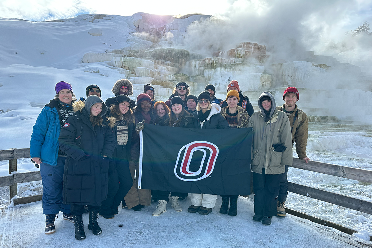 A group of UNO students stand in front of a geothermal feature at Yellowstone National Park, while holding up the UNO flag.