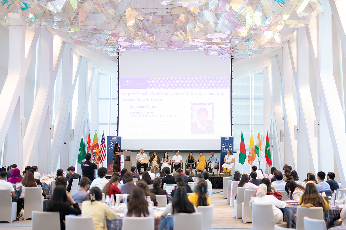 A crowd sits looking on at a panel of speakers sitting on a stage in a hotel conference room.