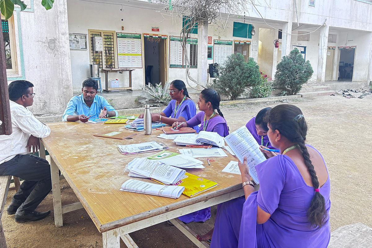 Adults sit around a table outside in a school courtyard reading curriculum.