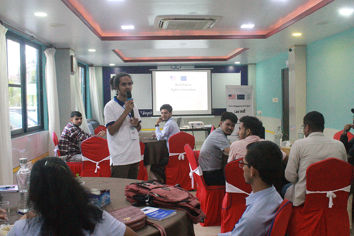 A young man holding a microphone speaks to others seated at tables around him.