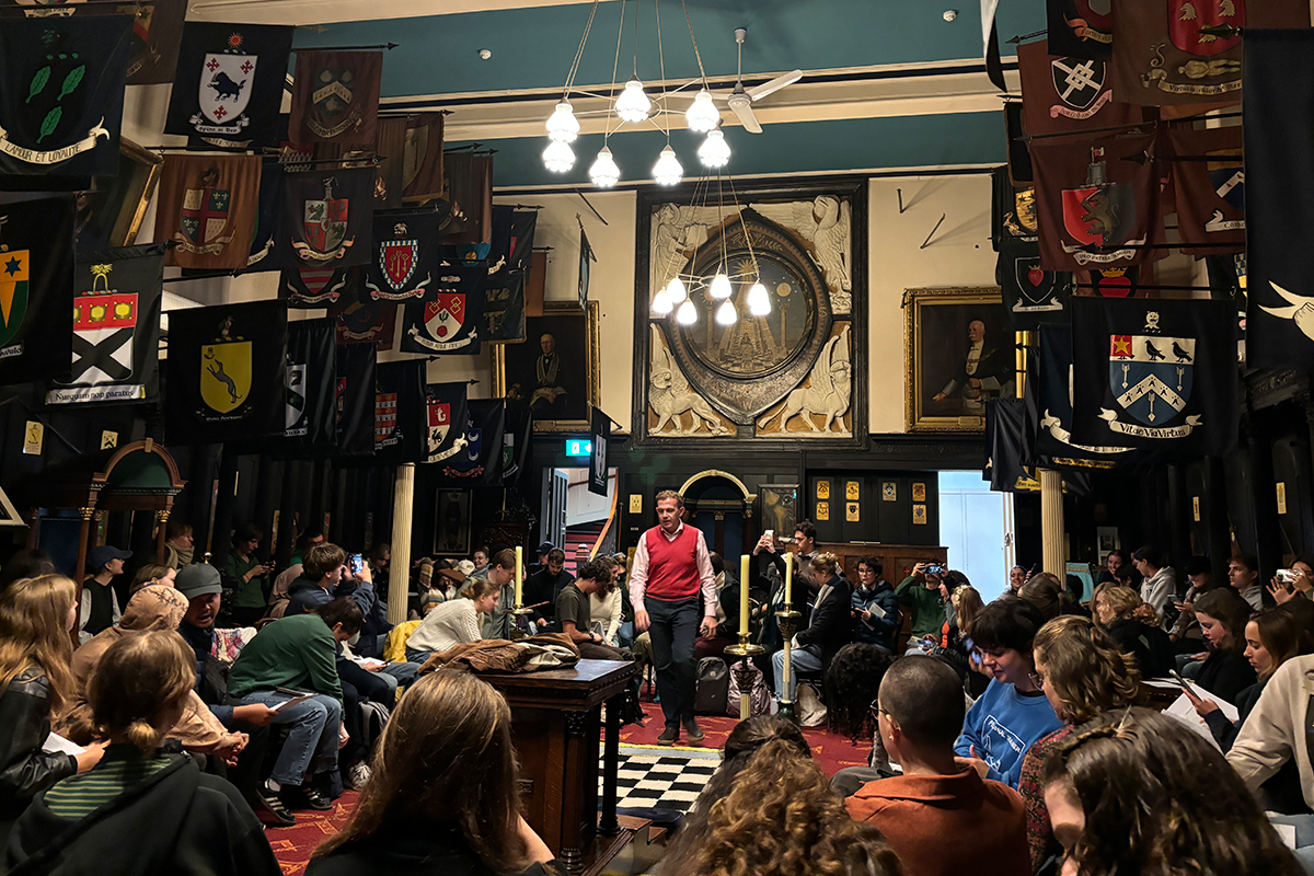 Students sit around a professor in an old style medieval hall with banners hanging from the ceiling.