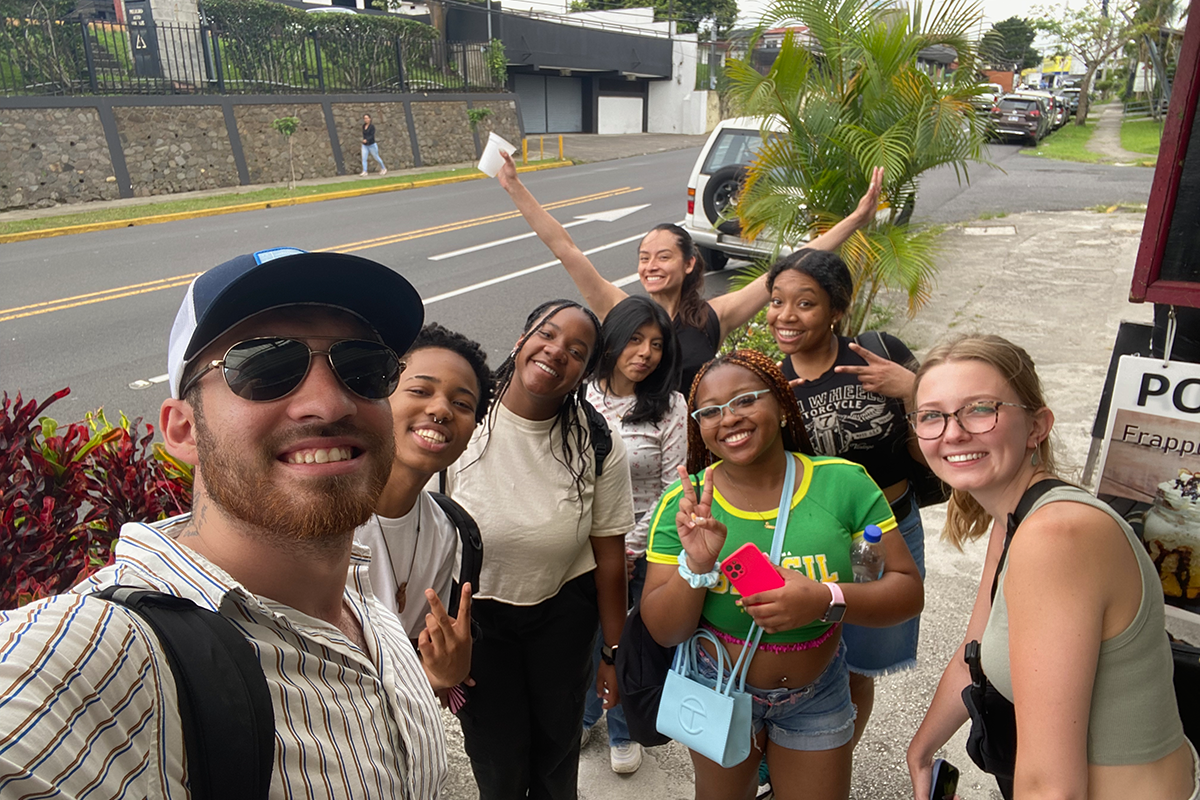Group of young adults pose together for a photo, smiling, with a road, cars, and palm plants in the background.