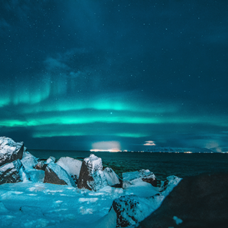 Snow covers a rocky beach overlooking the water, with the northern lights in the sky beyond.