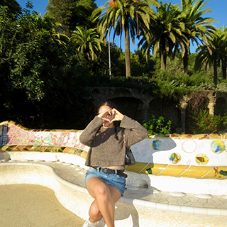 Young woman makes a heart shape with her hands while sitting on a mosaic lined bench with palm trees in the background.