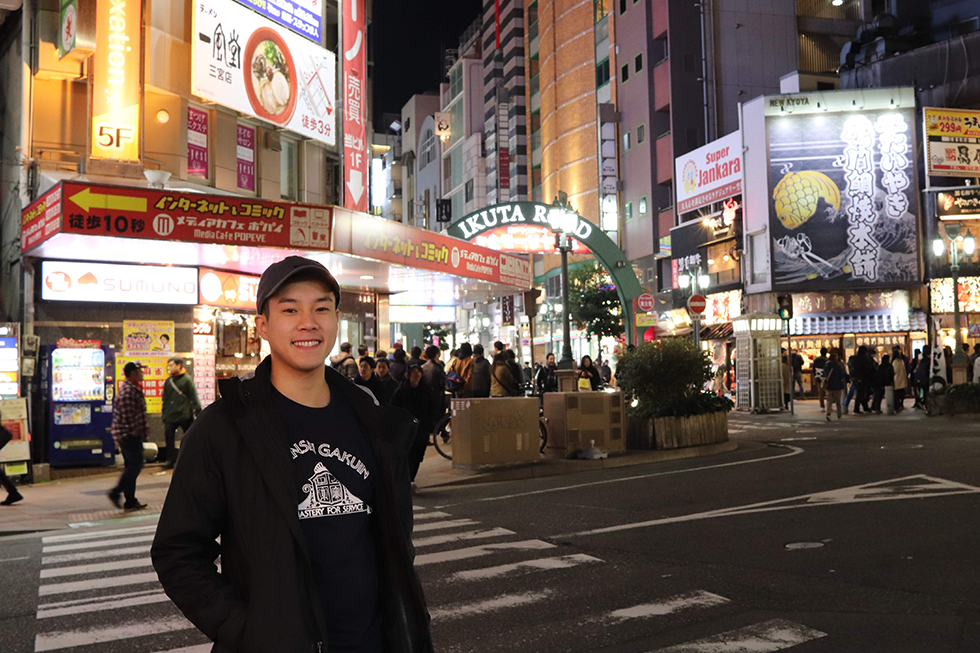 Young man in a baseball cap stands outside in a Japanese city at night.