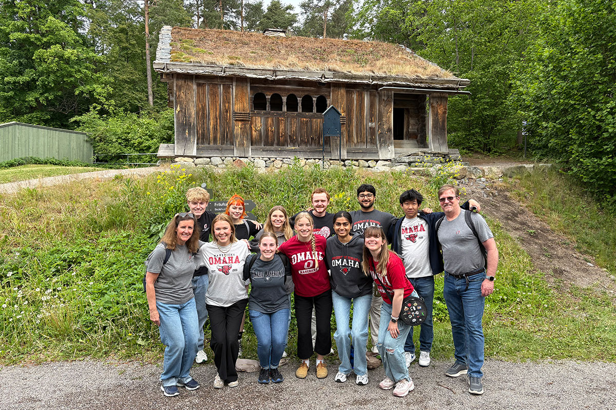 A group in University of Nebraska Omaha shirts poses outside in front of a small wooden building with a grass roof.