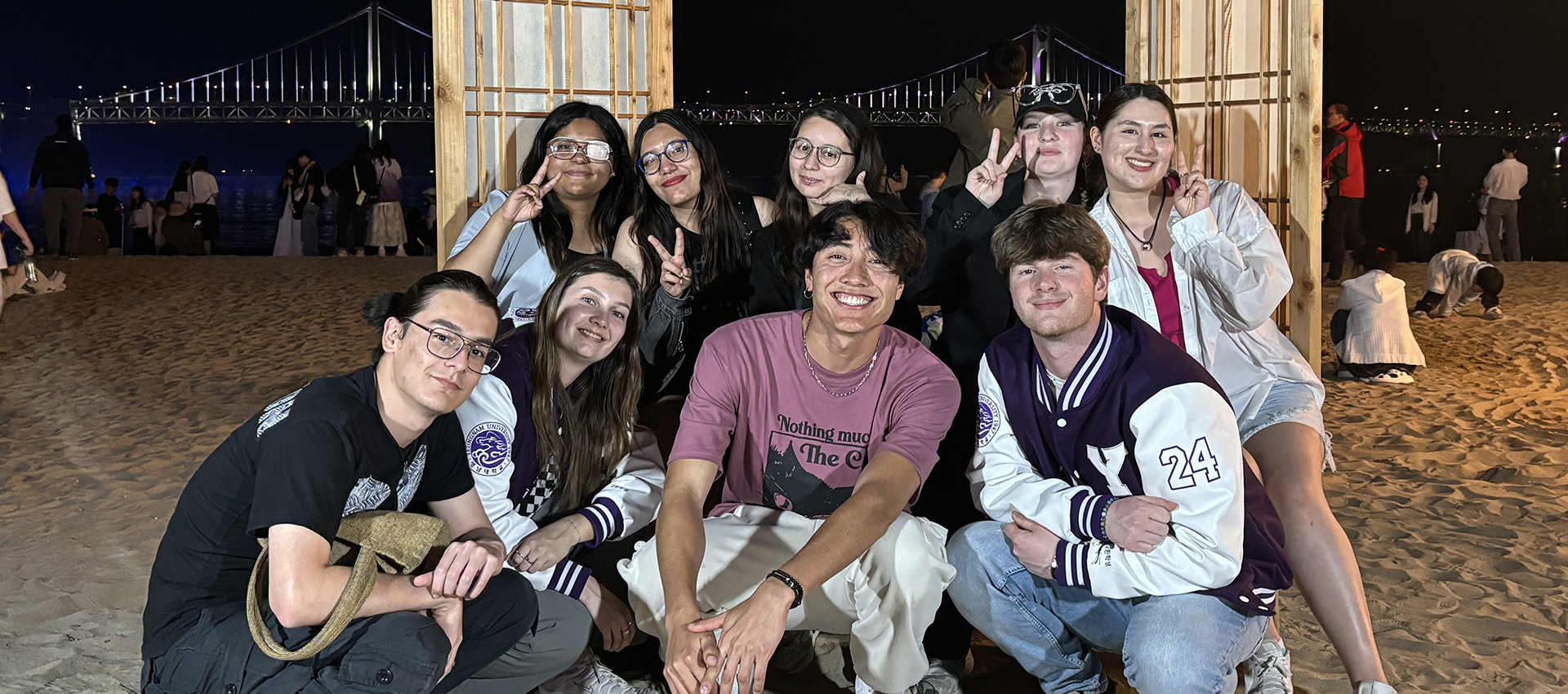 group of college students post for a photo on a beach in South Korea at night with a suspension bridge behind them.