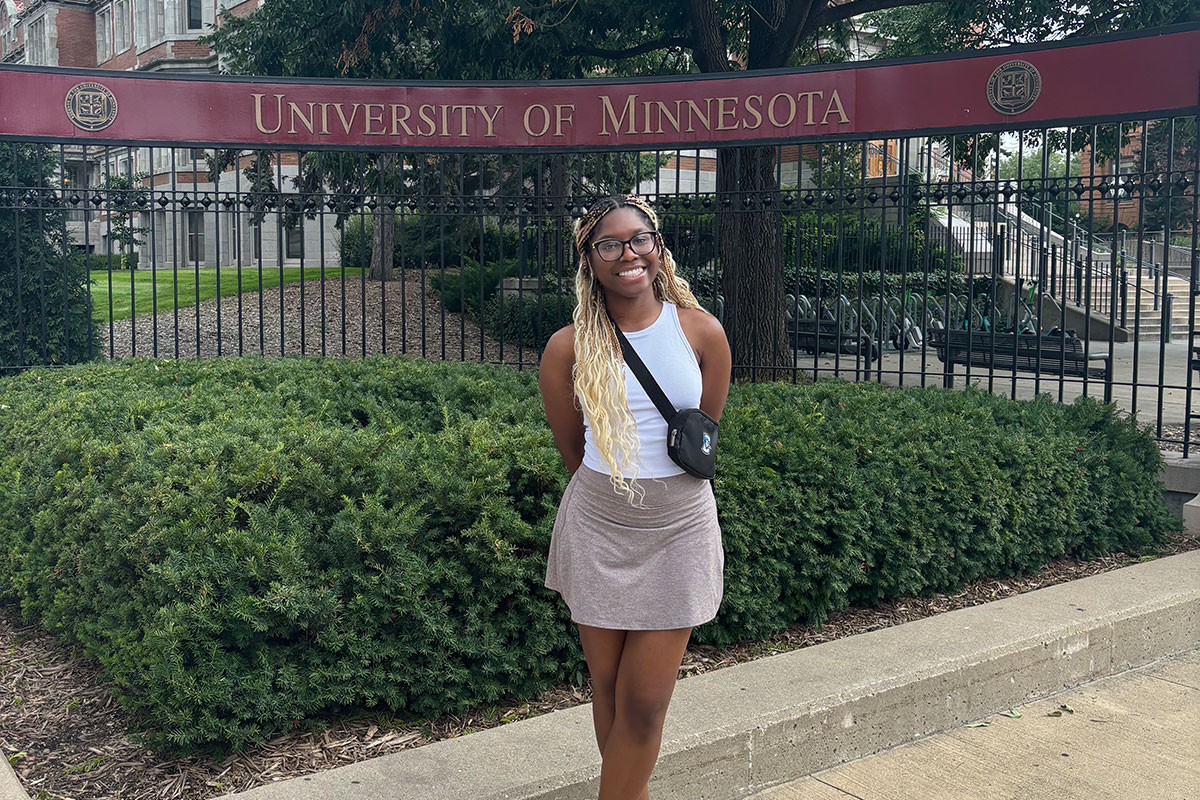 A female college student stands in front of a University of Minnesota sign outside.