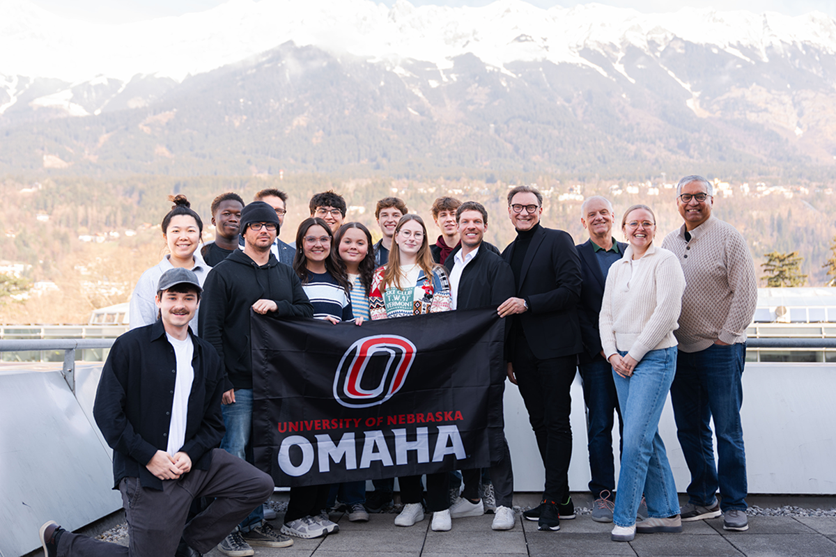 Group poses with University of Nebraska Omaha flag with snowcapped mountains in the background.