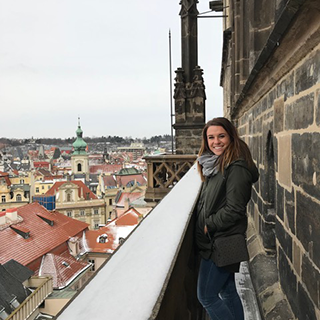 Young woman stands on a narrow balcony overlooking a European city.