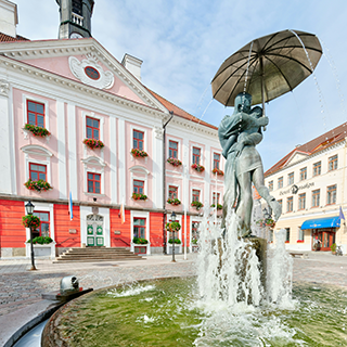 A large water fountain featuring a statue of a couple kissing under an umbrella is in front of a pink and white building in a public square.