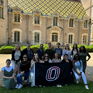 A group of college students pose for a photo with a University of Nebraska Omaha flag in front of a historic building in France.