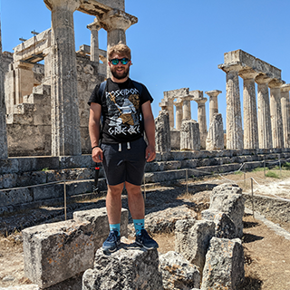Young man stands on a broken column surrounded by ancient Greek ruins.
