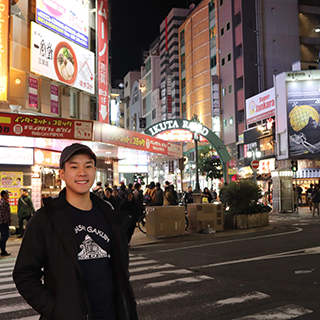 Young man stands in front of a busy street with people shopping at night.