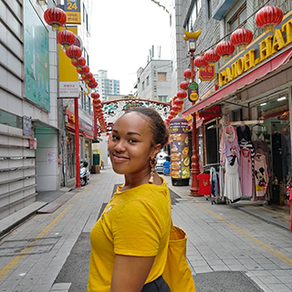 A young woman stands in the street looking at the camera over her shoulder, with shops and hanging red lanterns on either side.