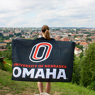 Young woman with back to camera holds a University of Nebraska Omaha flag over her shoulders while looking out at a city.